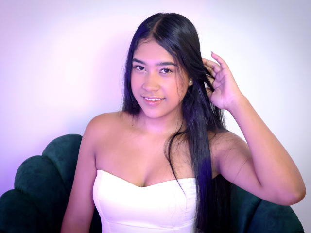 Ivonne Laurent, con cabello negro largo, vestida con un vestido blanco sin tirantes, sonriendo, con la mano en el cabello, silla de terciopelo verde, iluminación en tonos rosa claro y morado, linda, pechos pequeños, piel bronceada..