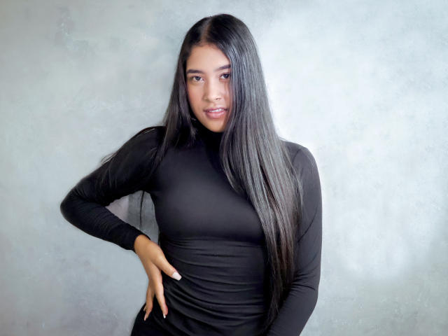 Ivonne Laurent, con cabello largo y negro, vestida con un vestido negro ajustado de manga larga, de pie frente a una pared gris, con una mano en la cadera, sonriendo suavemente y mirando directamente a la cámara..
