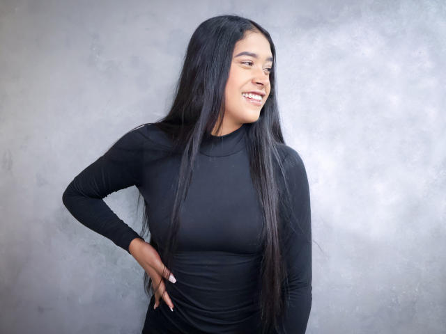 Ivonne Laurent, con cabello negro largo, sonriendo, vestida con una camiseta negra ajustada de manga larga, con las manos en las caderas, de pie frente a una pared gris con textura, mostrando sus pequeños senos y su cara linda..