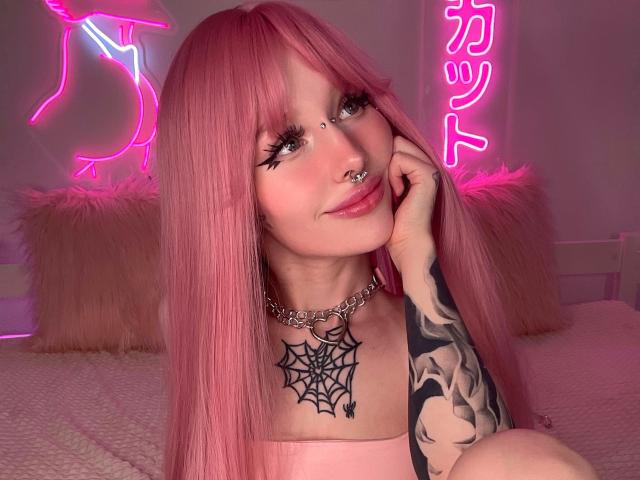 LaneyvilI with pink hair, septum piercing, spider web tattoo, tattooed arm, chain necklace, pink neon lights, and fluffy pillows, smiling softly. Boobs visible under pink top.