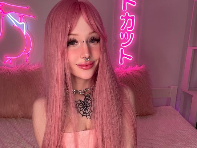 LaneyvilI, con cabello rosa, aro en la nariz y un collar con forma de telaraña, sonriendo en una habitación iluminada con luces de neón, vestida con un top rosa sin tirantes, mostrando sus pechos..