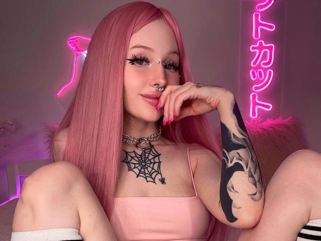 LaneyvilI, con cabello rosa, piercing en la nariz y un brazo tatuado, vestida con un top rosa de tirantes finos, sentada con las rodillas levantadas, mostrando sus pechos y un tatuaje de telaraña. Un letrero de neón en el fondo..