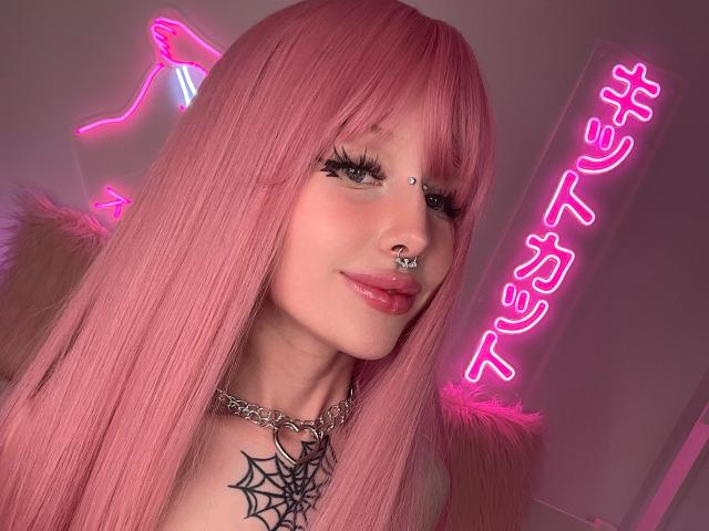 LaneyvilI with pink hair, green eyes, nose ring, and lip ring, wearing a chain necklace and spider web tattoo, neon pink background, and fluffy pink fur collar.