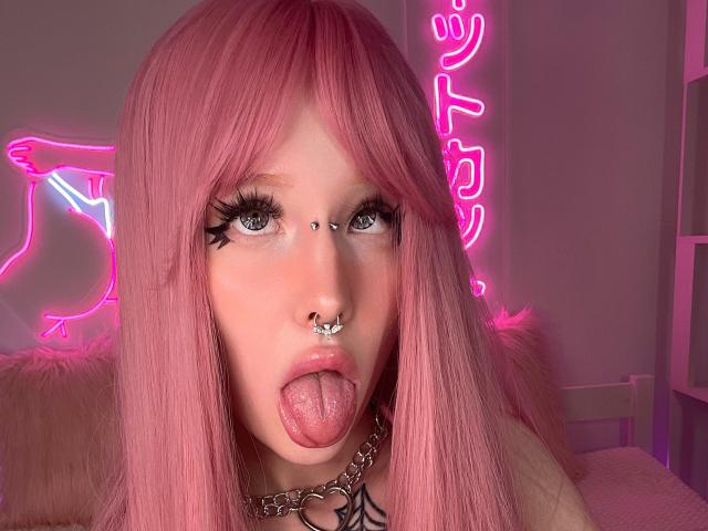 LaneyvilI con cabello rosa, ojos verdes, delineador de ojos negro, piercing en la nariz, piercing en el tabique nasal, piercing en el labio, lengua afuera, usando un collar de cadena, luces rosa neón en el fondo..