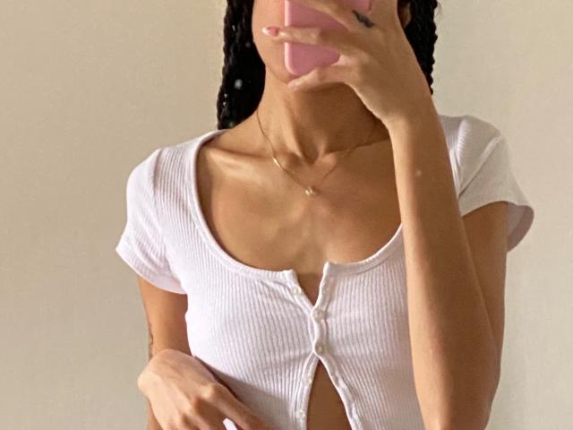 IrinaDrew with tan skin, braided hair, holding pink phone, wearing white ribbed button-up top, showing small tits, slim, gold necklace, taking mirror selfie.