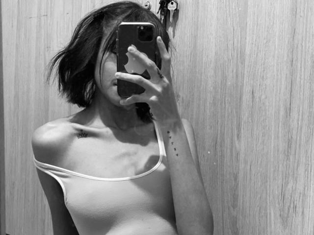IrinaDrew taking a mirror selfie, her phone covering her face. She's wearing a white off-shoulder top, showing small tits and a skinny, ribbed frame. Wood background.