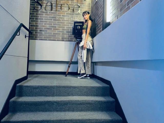 IrinaDrew stands on carpeted stairs, wearing a green tank top, gray pants, and black-and-white sneakers, holding a white shirt, with a vintage camera on a tripod.