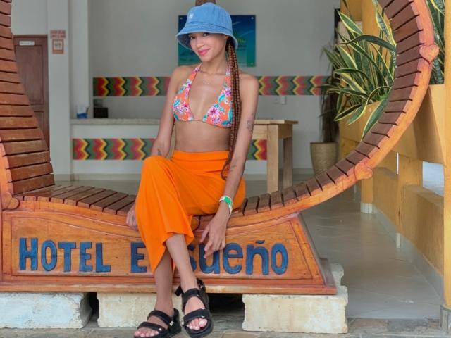 IrinaDrew with colorful bikini top, orange skirt, bucket hat, and black sandals, sitting on wooden bench, long braid, pink lipstick, green wristband, showing her tits and ass.