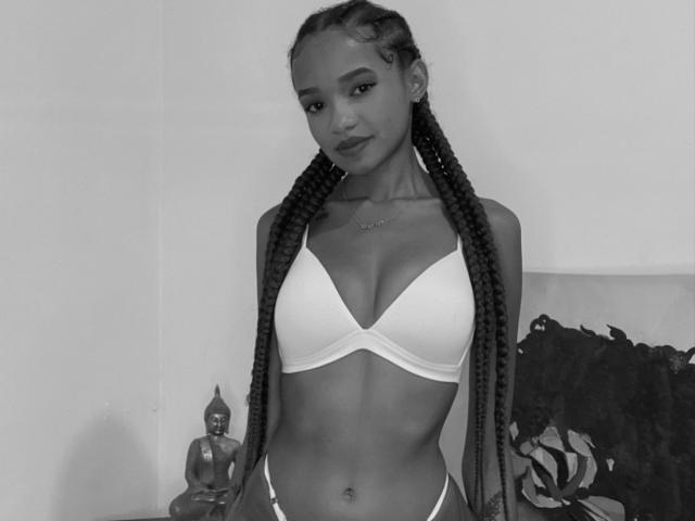 Black-and-white photo of IrinaDrew with long braids, small tits in a white bra, wearing a thong, standing, smiling, small belly button, Buddha statue on left, pillow on right.