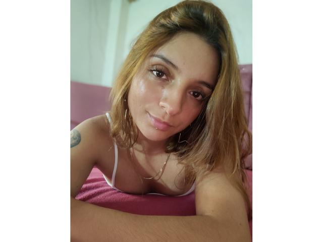 Saarah Jones, met lichtbruin haar, lichtjes open lippen, een subtiele glimlach, draagt een witte tanktop, waarbij haar kleine borsten zichtbaar zijn, liggend op een roze bed, met een tatoeage op haar rechter schouder, zacht licht..