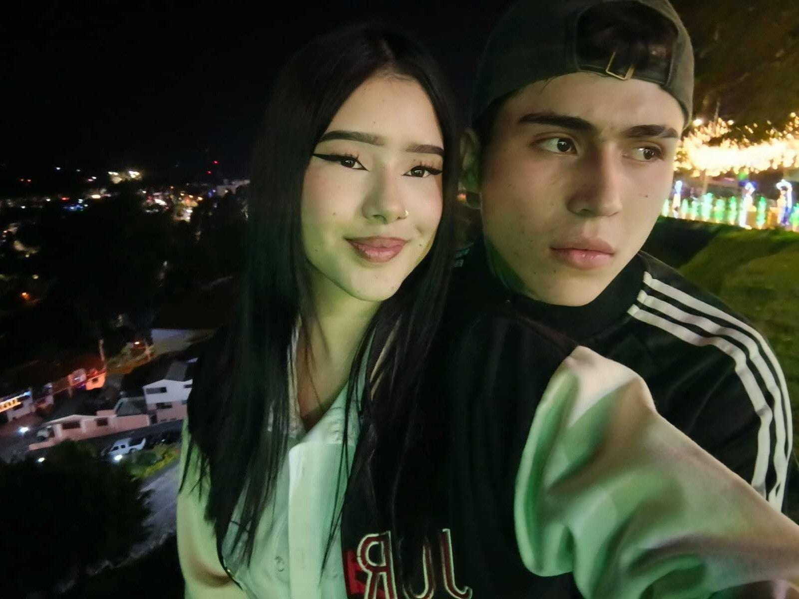 HinatayPhilip, a cute Asian girl with long black hair and small tits, stands next to a young dude with a baseball cap and athletic jacket, both smiling at night.