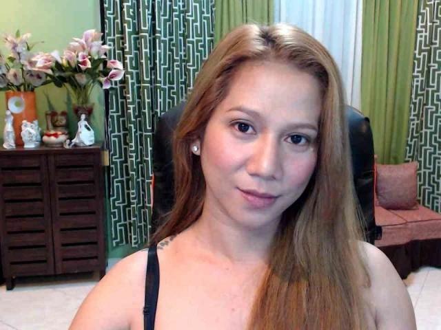 Katycumloader: atractiva, con cabello rojo largo, un top negro y maquillaje discreto, sonriendo ligeramente, mostrando pequeños senos y un rostro lindo, en una habitación con cortinas de color verde con estampado..