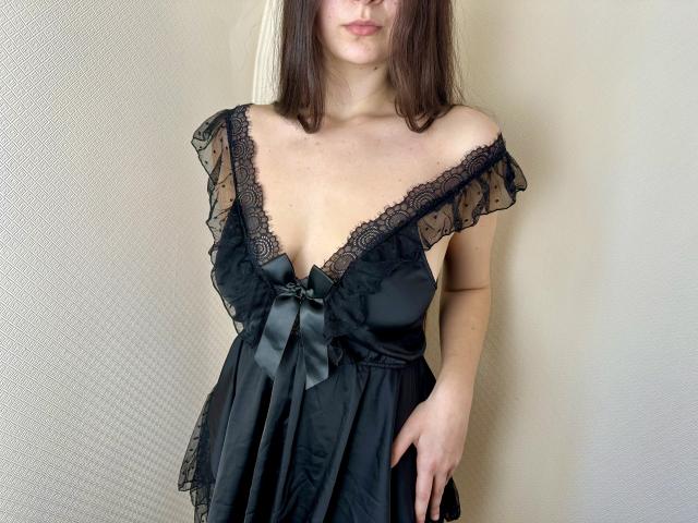 Tisia with medium tits, wearing a black, lace-trimmed, off-shoulder nightie, has a black satin bow on her chest, and her right hand in the dress's skirt.