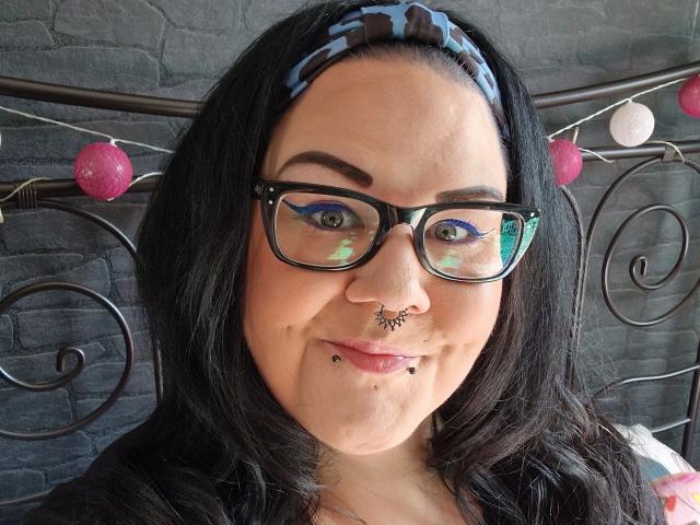 Scharfe-kitty ha i capelli neri, una fascia per capelli blu con disegni, occhiali neri, un anello al naso, un piercing al setto nasale, un anello al labbro e un eyeliner colorato. Il seno non è visibile. Sorride leggermente..