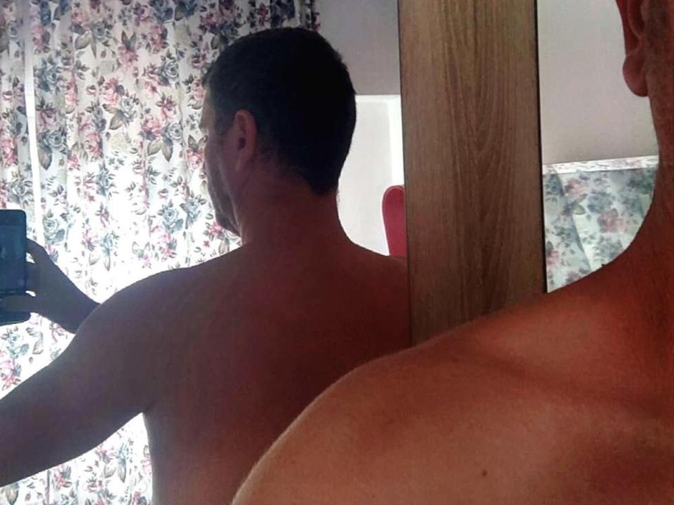 Een naakte man met kort bruin haar, staand met zijn rug naar een spiegel, die een foto maakt van zijn reflectie. Zijn blote billen en rug zijn zichtbaar..