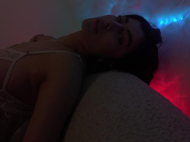 EmmaBeaumont lying on a couch, head tilted back, dark hair, white lace bra, dim red and blue light, relaxed expression, soft gray cushion, subtle shadow, sensual mood.
