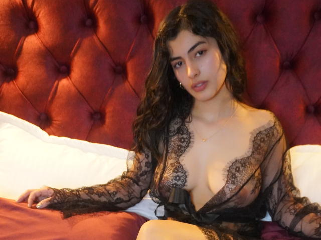 EmmaBeaumont with curly black hair, wearing a sheer black lace robe, exposing her boobs, sitting on a bed with a red tufted headboard and white pillows.