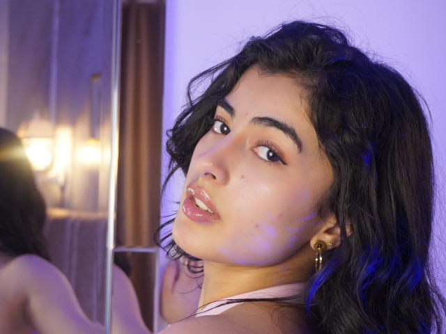 EmmaBeaumont with wavy black hair, full lips, and brown eyes looking over her shoulder. She's wearing a gold earring, and her pink halter top is visible. Soft purple lighting.