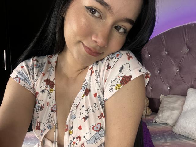 DeisyWest with long black hair, wearing a floral dress, showing her tits, smiling softly, in a bedroom with a purple tufted headboard. Her makeup includes winged eyeliner.