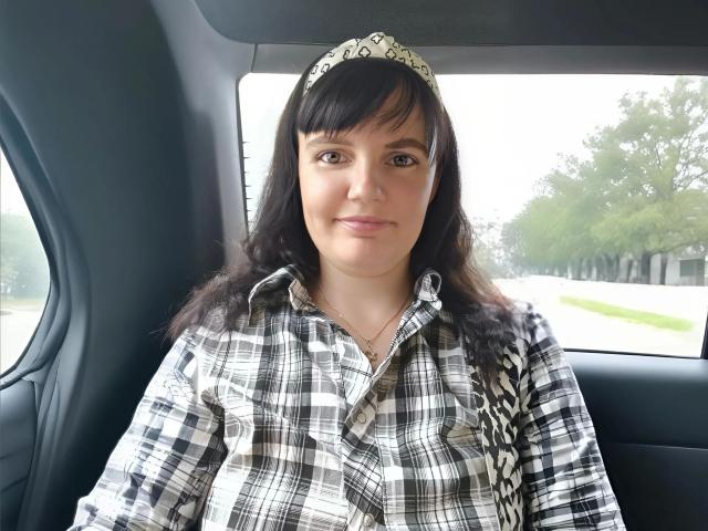 Alicexbaby69-hot has black hair, brown eyes, and wears a silver tiara with a plaid shirt. She's sitting in a car, smiling, with a black and white pattern.