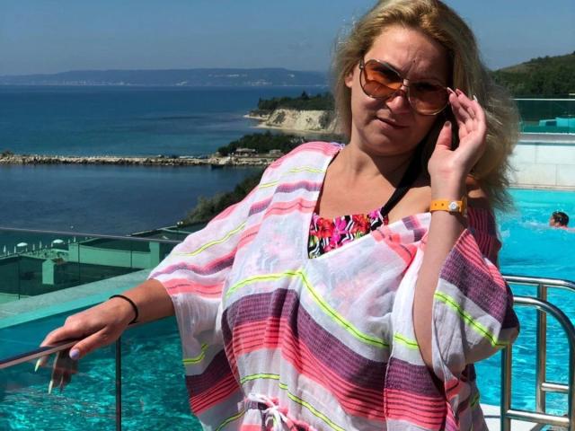 lisabigtits-hot with blonde hair, wearing orange sunglasses, pink and white striped shawl, and colorful top, leaning on pool railing, talking on phone, by blue ocean, sunny.