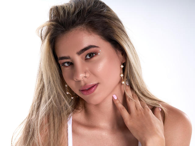 RileySwet has long blonde hair, brown eyes, and glossy pink lips. She's wearing white earrings with pearls and has her hand near her neck, showing pink nails.
