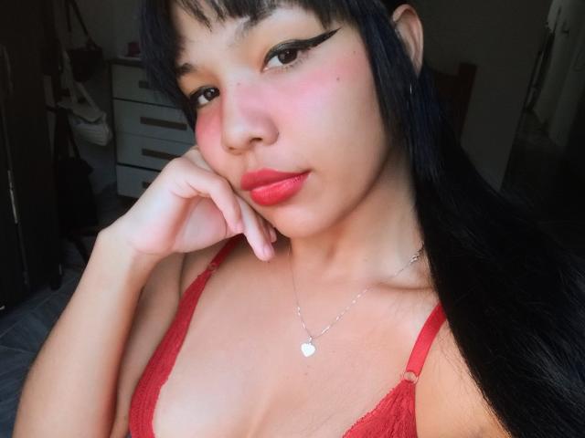 JadySousa with red lace bra, pink blush, black eyeliner, red lipstick, heart necklace, black hair, touching cheek, medium tits, light brown skin, bedroom background, confident expression.