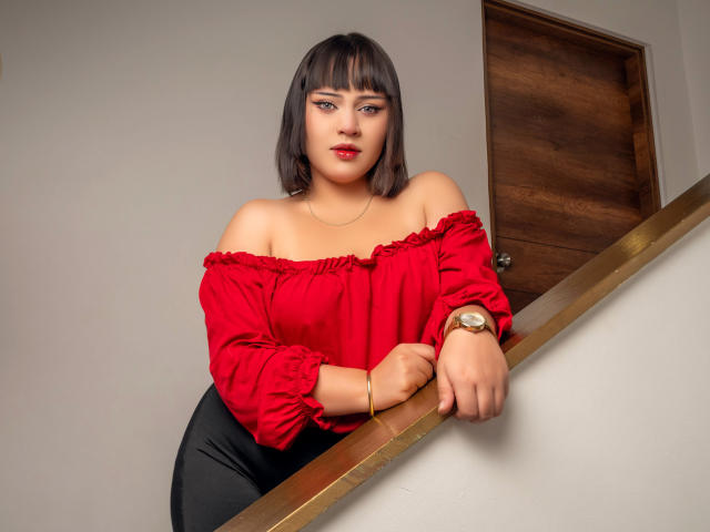 NymeriaMars with shoulder-length brown hair, red off-shoulder top, black pants, and gold watch, leaning on a wooden staircase railing, looking at the camera with red lipstick.