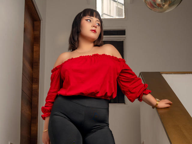 NymeriaMars has short black hair, red off-shoulder top, black high-waisted pants, and a gold bracelet, standing with one arm on a wooden railing, looking up with a slight smile.