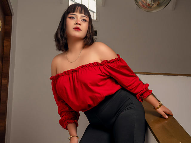 NymeriaMars with medium tits, wearing a red off-shoulder top and black pants, leaning on a wooden railing, short brown hair, red lipstick, gold bracelets, looking up.