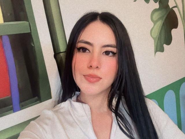 EimmyPark, con lunghi capelli neri, pelle chiara e eyeliner allungato, indossa una giacca bianca con zip, sorride dolcemente, guardando la fotocamera, seduta in una stanza verde e bianca..