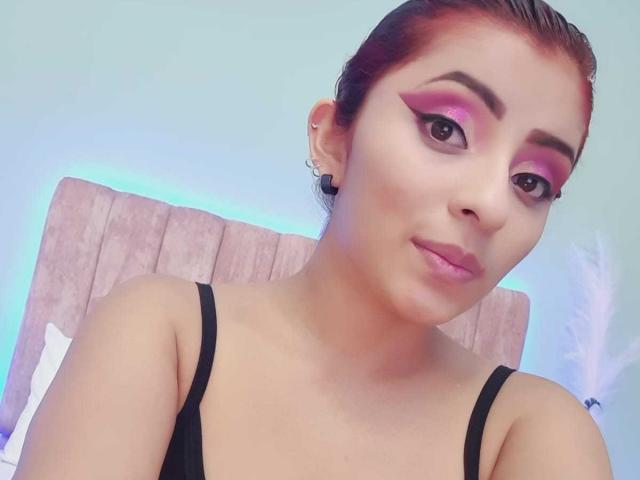 LizBonnet trägt rosigen Lidschatten, schwarzen Eyeliner und rosafarbenen Lippenstift, ein schwarzes Top mit Trägern, ihre braunen Haare sind zurückgebunden, und sie hat ein leichtes Lächeln, wobei kleine Ohrringe zu sehen sind..