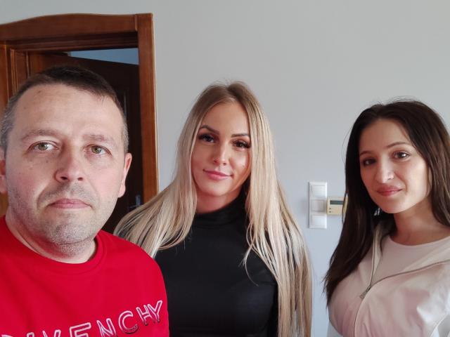 Drei Personen, Sophie69Trio mit kurzen, grauen Haaren in einem roten Hemd, eine blonde Frau in einem schwarzen Rollkragenpullover und eine braunhaarige Frau in einem weißen Hemd, stehen zusammen und lächeln in die Kamera..