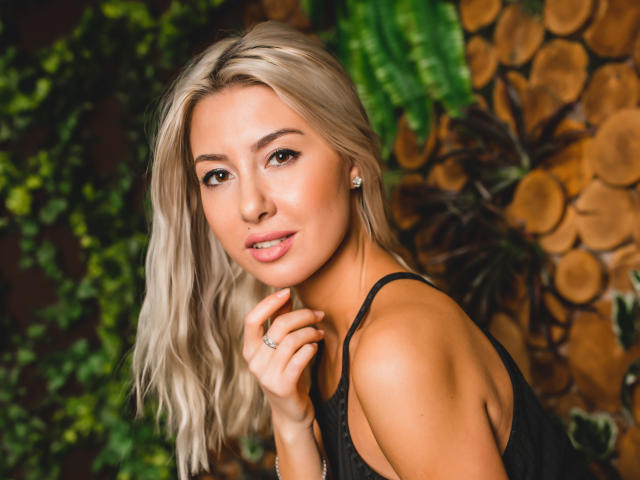 VictoriaRay with blonde hair, cute face, and small tits, wearing a black top, looking at camera, hand near chin, green plant and wood background. She's smiling softly.