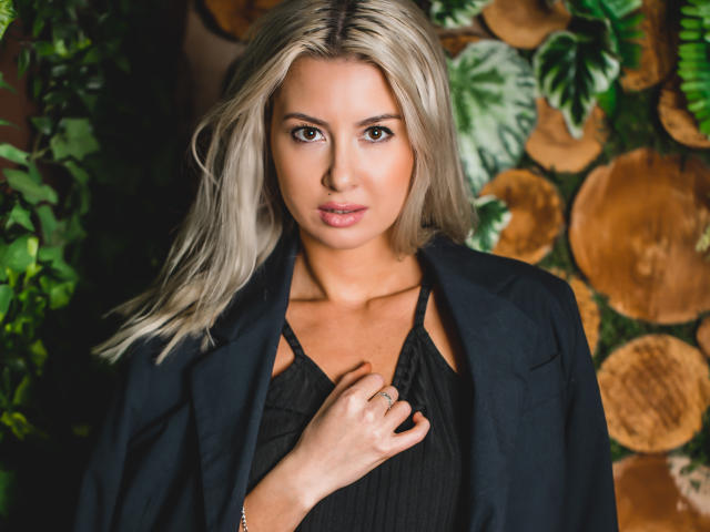 VictoriaRay with blonde hair, black dress, and blazer, holding her chest, showing her tits, against a wooden log wall with green leaves. Her brown eyes are focused.