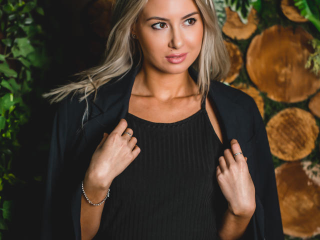 Victoria Ray mit blonden Haaren, die ein schwarzes Kleid und einen offenen schwarzen Mantel trägt, die Ränder des Mantels hält, ein Armband am rechten Handgelenk, dezentes Make-up, blickt in die Kamera, kleiner Nasenring..