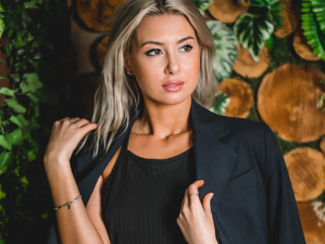 VictoriaRay with blonde hair, wearing a black top and jacket, holding jacket collar, looking to the side, subtle makeup, bracelet on left wrist, ring on right hand, attractive.