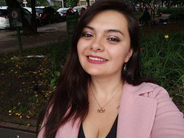 KatherinLatin with long brown hair, pink coat, black top, showing her boobs, smiling, pink lipstick, nose ring, gold necklace, standing in a park with green grass.