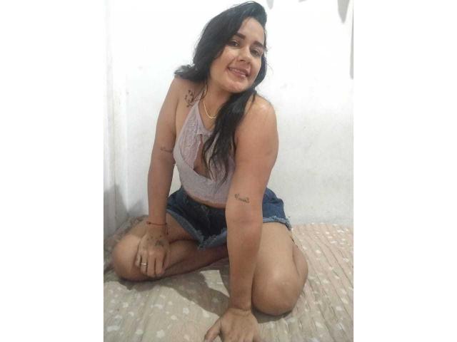 ScarletJamiesson, with long black hair, wearing a purple lace top and denim shorts, is kneeling on a patterned floor, smiling, showing tattoos on her arms and a gold necklace.
