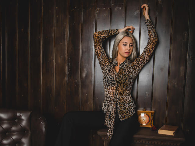 VictoriaRay with blonde hair, leopard print blouse, and black pants, arms raised, leaning against dark wooden wall, vintage clock on wooden table, brown leather couch in background.