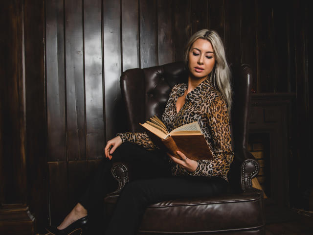 VictoriaRay with blonde hair, leopard print shirt, black pants, reading on brown leather chair, holding open book, looking down, in dimly lit, wood-paneled room.