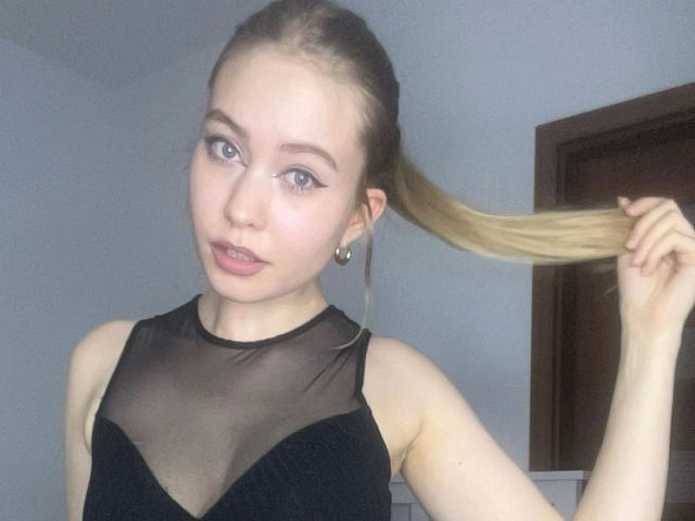 BrandyHelga-hot with blonde hair, holding a ponytail, wearing a black sheer top, showing small tits, pale skin, blue eyes, and pink lips, in a simple room.