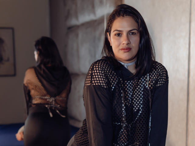 ElizabethDupont with dark brown hair, wearing a black mesh top, looking at the camera, with her back to the mirror showing her ass. She has a serious expression.