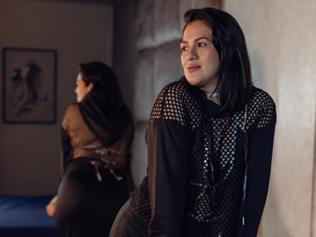 ElizabethDupont with black mesh top, showing her ass in reflection, black hair, looking over shoulder, soft smile, dark room, blue bed in background, mirror on wall.