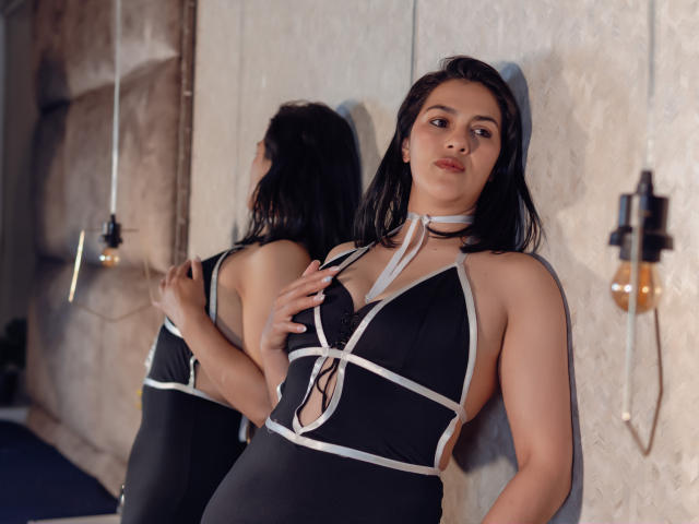 Elizabeth Dupont, vêtue d'une robe noire et blanche à bretelles, dévoilant ses seins dans une robe à lacets, se tient devant un mur avec un miroir, qui reflète ses fesses, dans une pièce faiblement éclairée..
