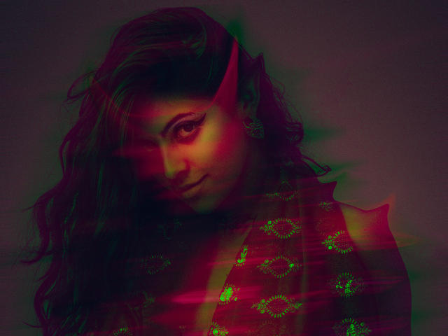 CoriPaige's face with red and green light effects, wavy dark hair, and a subtle smile, showing a slightly melancholic expression, with a patterned red garment partially visible.