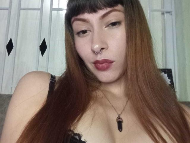 AmyLeeHot mit langen, braunen Haaren, Nasenring und rotem Lippenstift, die die Lippen leicht geöffnet zusammenpresst. Sie trägt eine schwarze Spitzenbluse und eine Halskette mit einem violetten Stein..