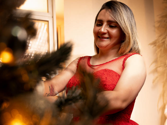 LiliEvanss in red lace dress, smiling, decorating a Christmas tree, her tits visible, blonde bob, tattoo on left arm, warm indoor light, tree lights in the foreground.