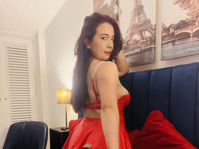 AnaGerald with red dress, showing her ass, sitting on blue bed, long dark hair, red lips, looking back, small earrings, room with lamp and Paris wall art.