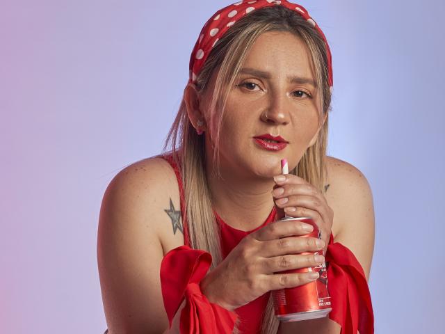 LiliEvanss with blonde hair, red polka dot headband, red dress, holding red lipstick, slightly parted lips, visible star tattoos on shoulders, light pink-blue gradient background.